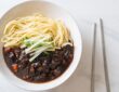 Seporsi jajangmyeon dengan saus kacang hitam yang kaya rasa, disajikan dengan acar lobak kuning dan mi gandum tebal, menciptakan hidangan khas Korea yang otentik