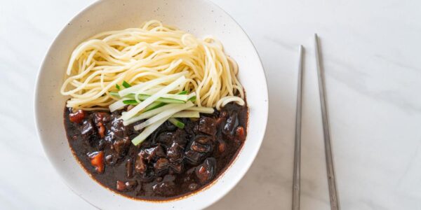 Seporsi jajangmyeon dengan saus kacang hitam yang kaya rasa, disajikan dengan acar lobak kuning dan mi gandum tebal, menciptakan hidangan khas Korea yang otentik